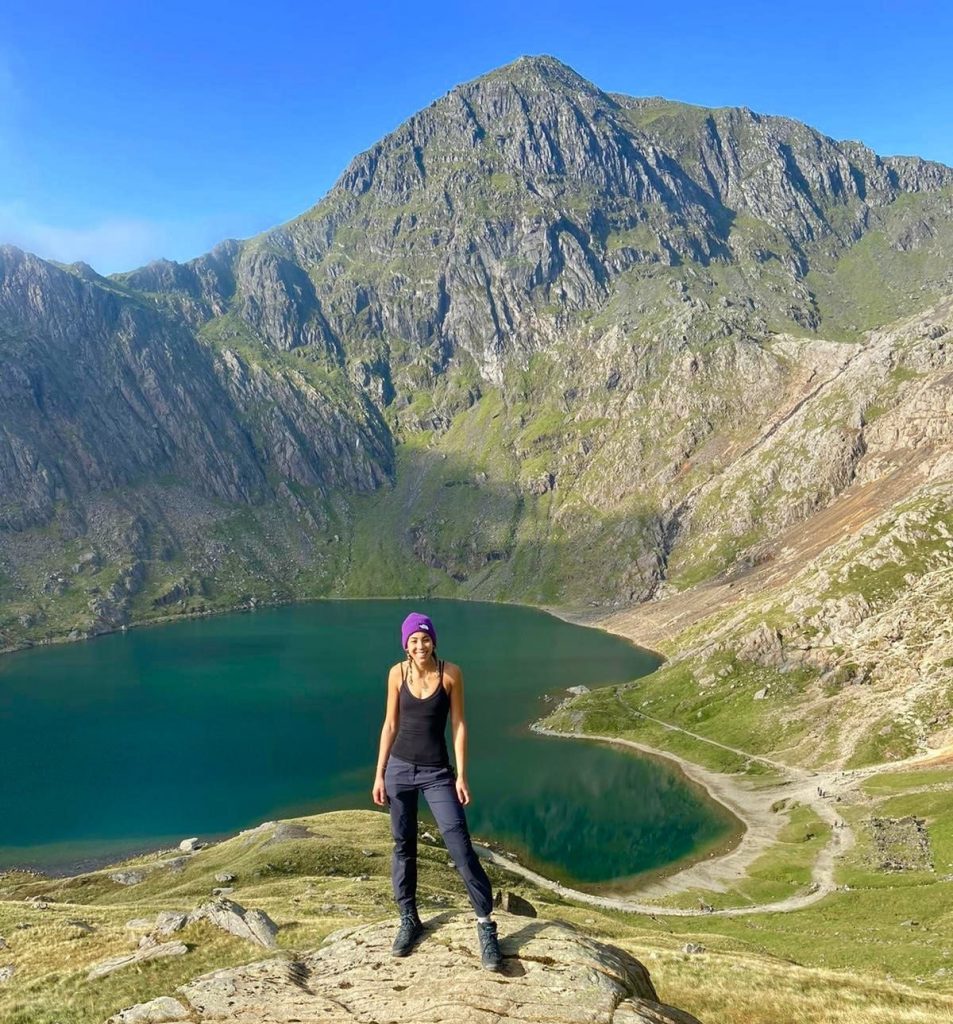Picture of Jade standing on a hill in the Lake District. There is a lake in the centre at the back with a mountain range surrounding it. Jade is a mixed race woman with medium-length brown and blonde hair. She is wearing black trousers and a black vest with a bright purple beanie. She is facing the camera with a smile.