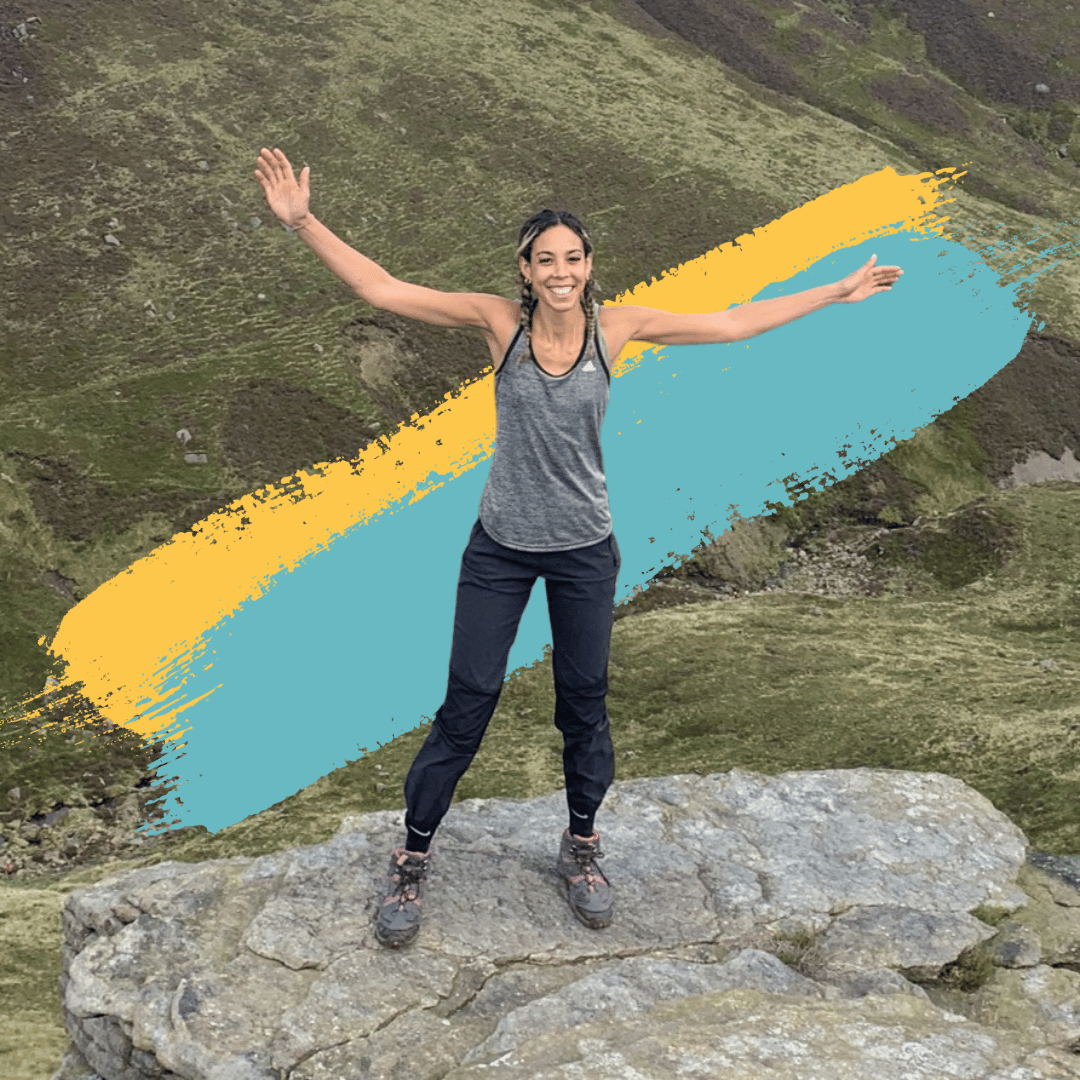 Picture of Jade Adams White standing on a hill in the lake district. She is facing the cameras with her arms wide in the air and a smile. Jade is a mixed race woman with medium length brown and blonde hair. She is wearing black trousers and a grey vest and her hair is in braids.