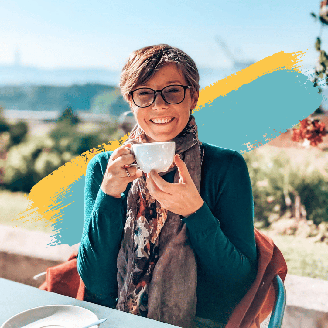 Picture of Joanna Horanin. She is a white woman with short auburn hair and glasses. She is sat down, holding a mug as she smiles to the camera. She is wearing a teal cardigan with a floral scarf. In the background there are light blue and yellow paint strokes.