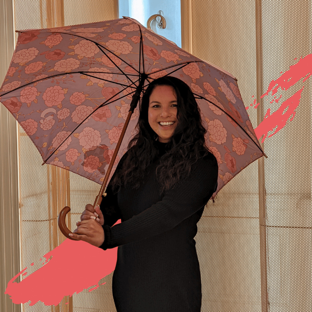 Picture of Rebekah Capon. Rebekah is a white woman with long dark brown hair. She is facing the camera while smiling and holding an umbrella. There is a horizontal graphic red brush stroke behind here.