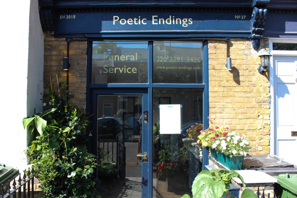 Picture of Poetic Endings funeral service office front. There is a small garden with big leafy plants and flowers outside. The wooden heading and wooden rims round the glass window and door is dark blue. "Poetic Endings" is a golden color.