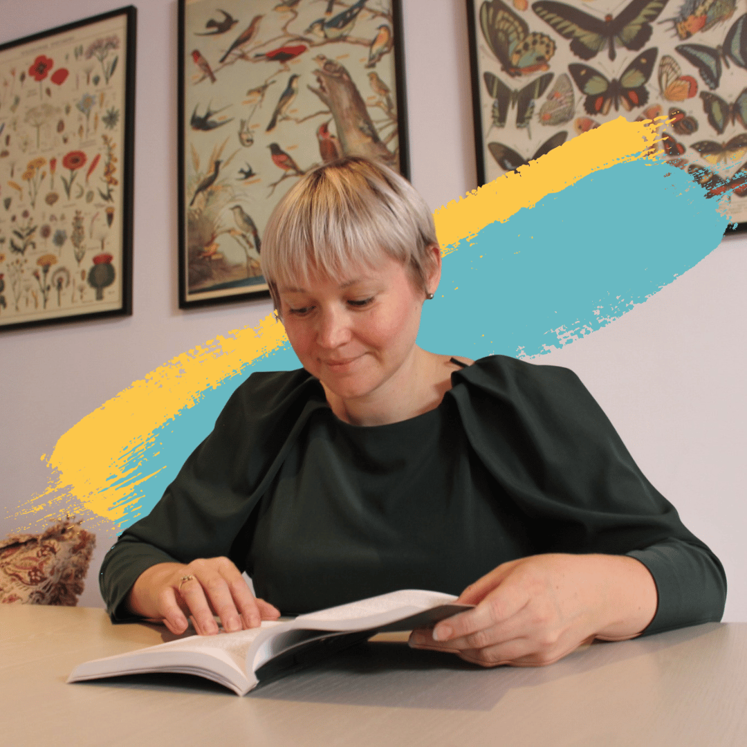 Picture of Louise Winters, smiling as she reads a book. Louise is a white women with a blonde pixie cut. She is wearing a dark green dress with puffed sleeves. In the background there are pictures or wildlife hanging on the wall. Behind her are blue and yellow graphic paint strokes.