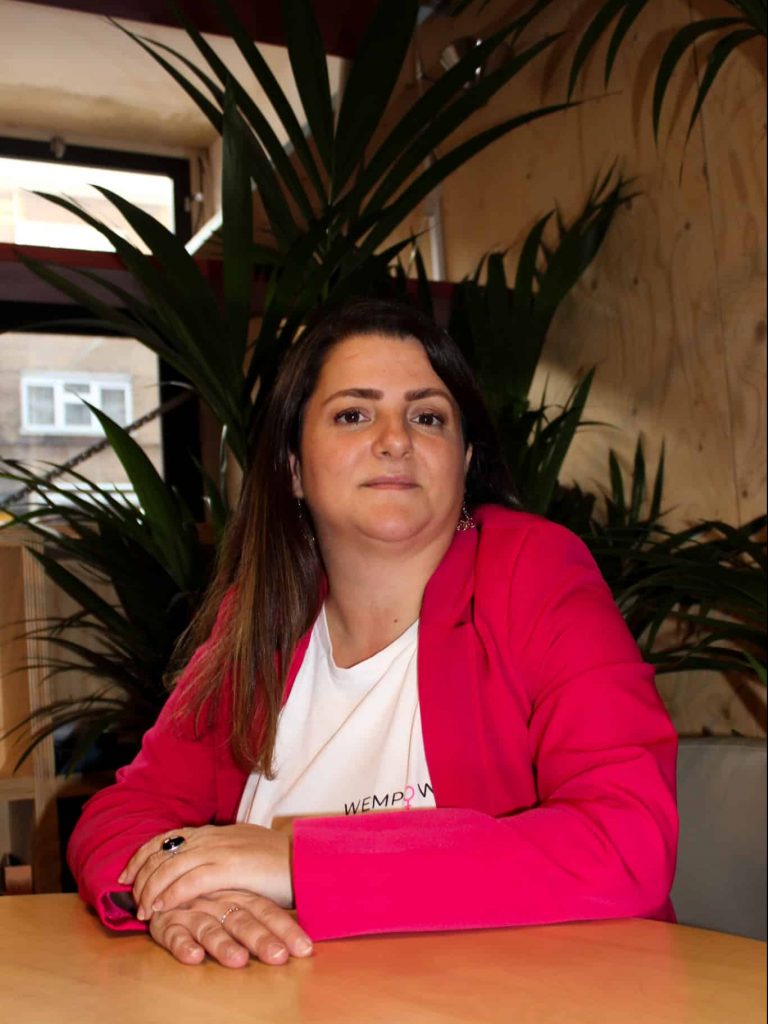 Picture of Ilaria Biancacci, the Hatch founder empowering women. Ilaria is a white woman with long brown hair. She is sat behind a table, with her arms folded over it as she faces the camera. She is wearing a hot pink suit jacked and a white shirt.