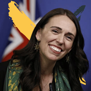 Picture of Jacinda Arden, former Prime Minister of New Zealand. Jacinda Arden is a white woman with long brown hair. She is looking at the camera with a smile, with the New Zealand flag in the background.