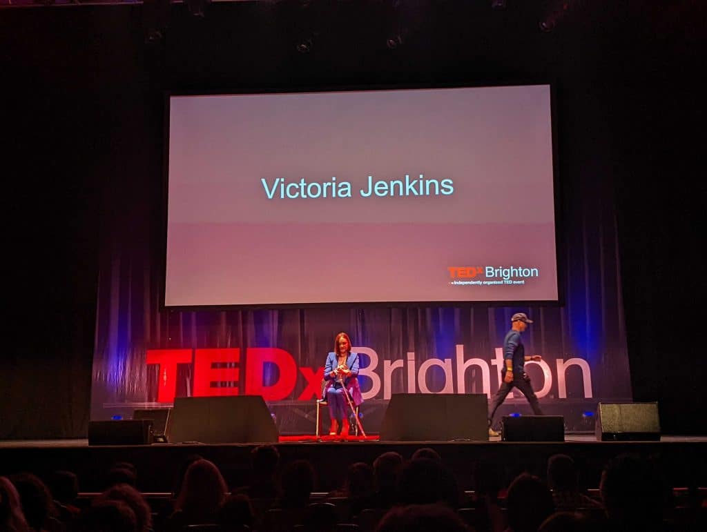 A woman with a walking stick is sat on a chair on a stage. There is a sign that says TED Brighton and the screen says Victoria Jenkins.
