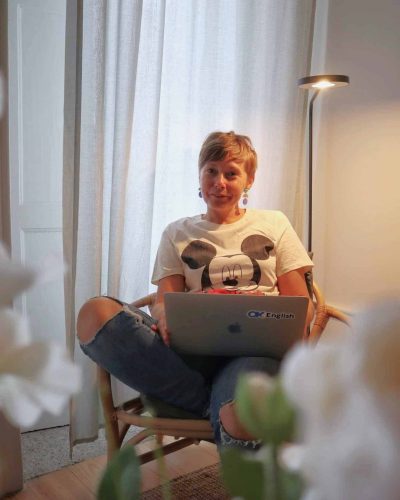 Picture of Joanna Horaning. She is sitting cross legged in a chair with a laptop in her lap. She is smiling at the camera. She is wearing ripped jeans and a Mickey Mouse t-shirt